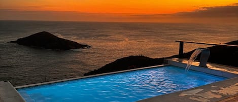 Una piscina con terraza de madera y vista al océano al atardecer.