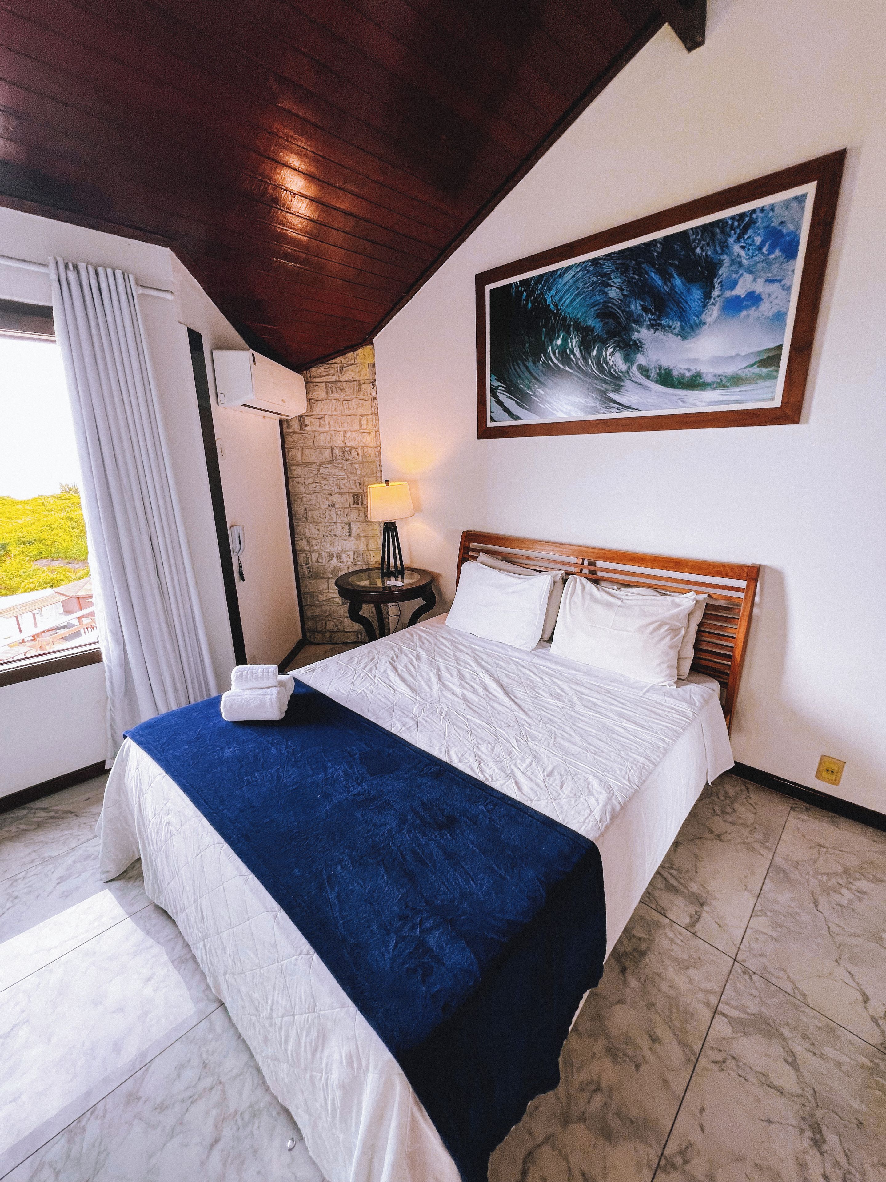 Habitación de hotel con una cama, un cuadro de una ola, una lámpara y vistas a zonas verdes al exterior.