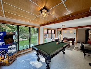 A pool table in a room with large windows, a foosball table, and a space-themed arcade machine.
