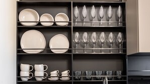 A kitchen with black shelves holding plates, bowls, cups, and glasses. There is a fruit bowl with bananas and apples on the counter. - ICONIC SALONICA SUITE seafront (Thessaloniki)