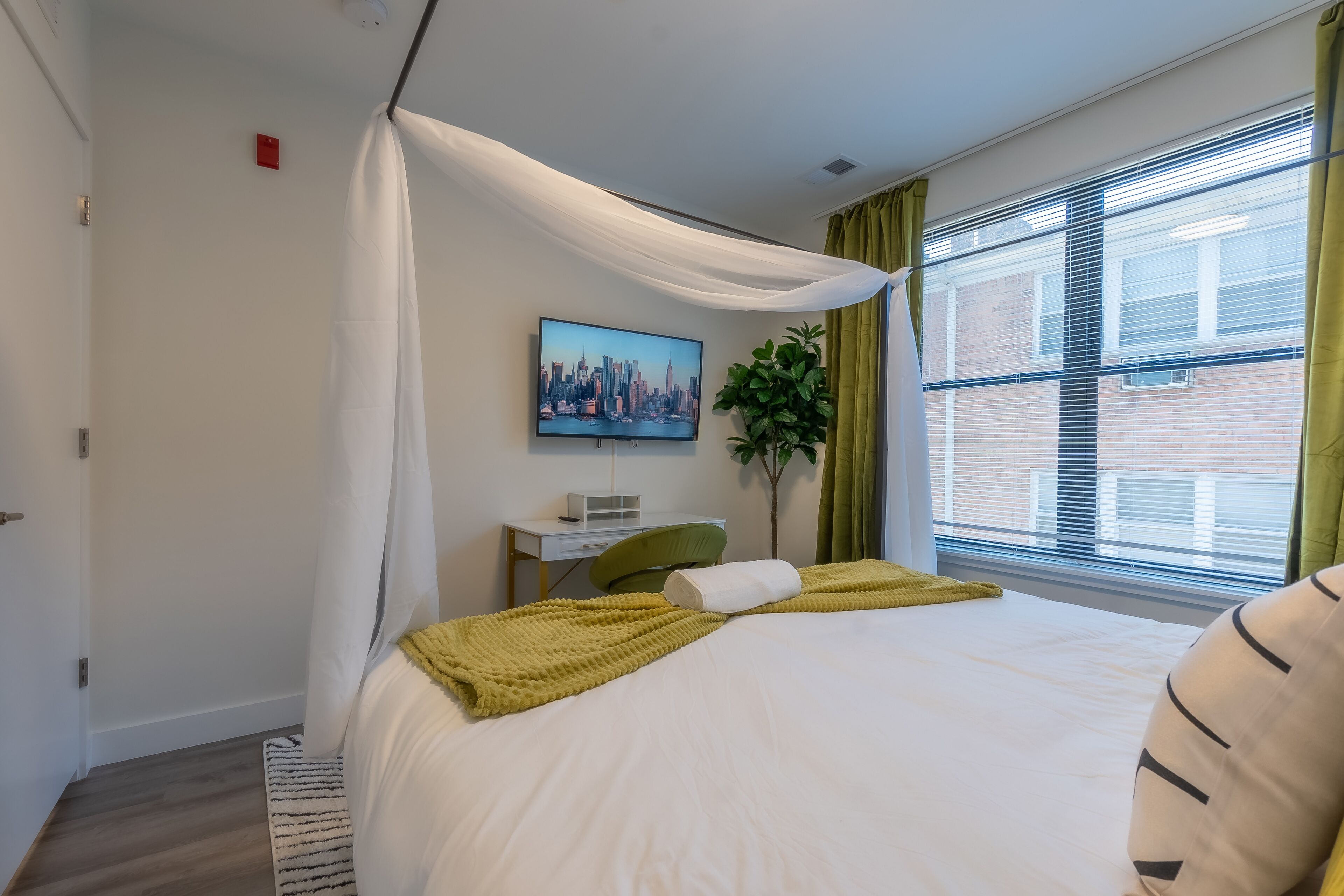 A hotel room with a bed, a desk, a chair, a TV, and a window with blinds.