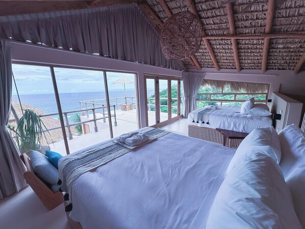 Quarto com uma cama grande, vista para o mar e um telhado de palha.