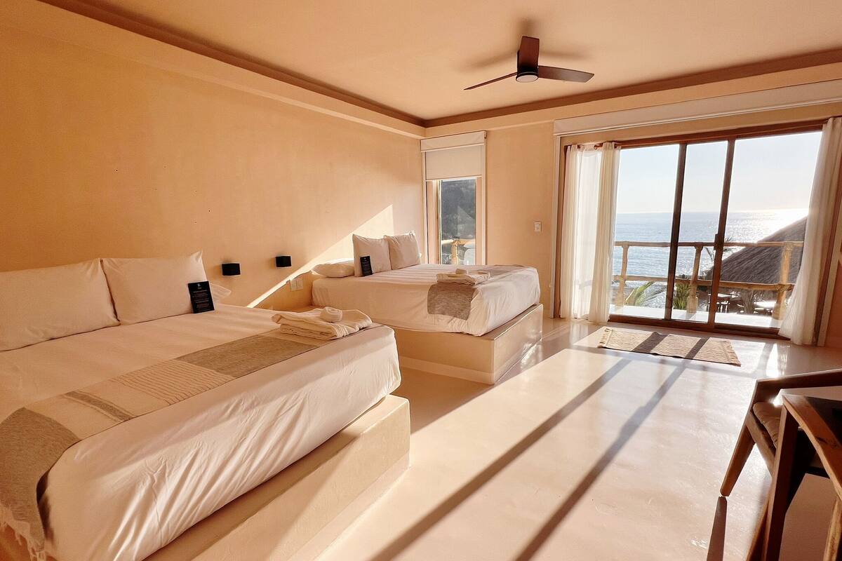 A hotel room with two beds, a ceiling fan, a large window with a view of the sea, and a balcony.