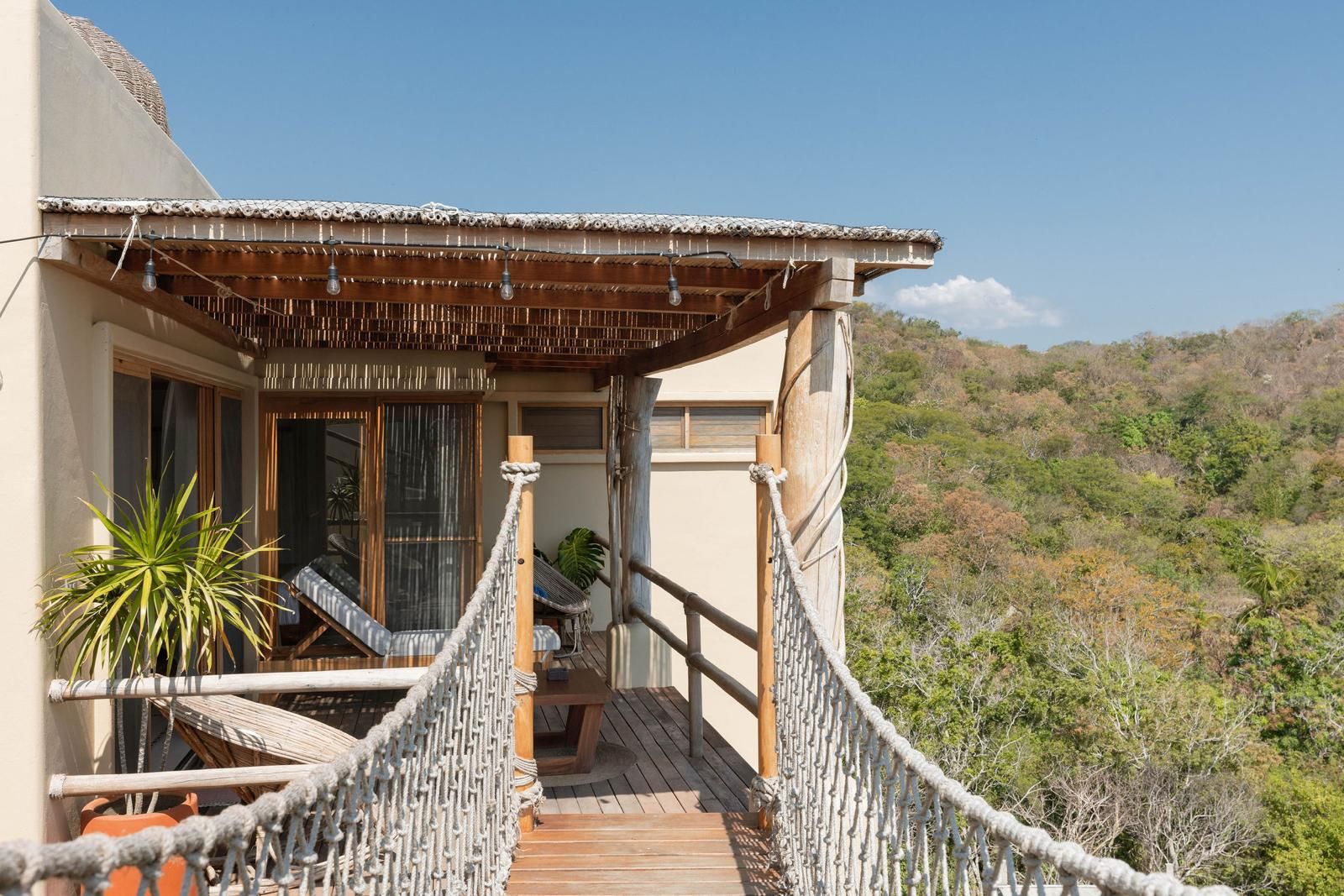 A wooden deck with a rope bridge leading to a covered seating area with a view of a hilly landscape.