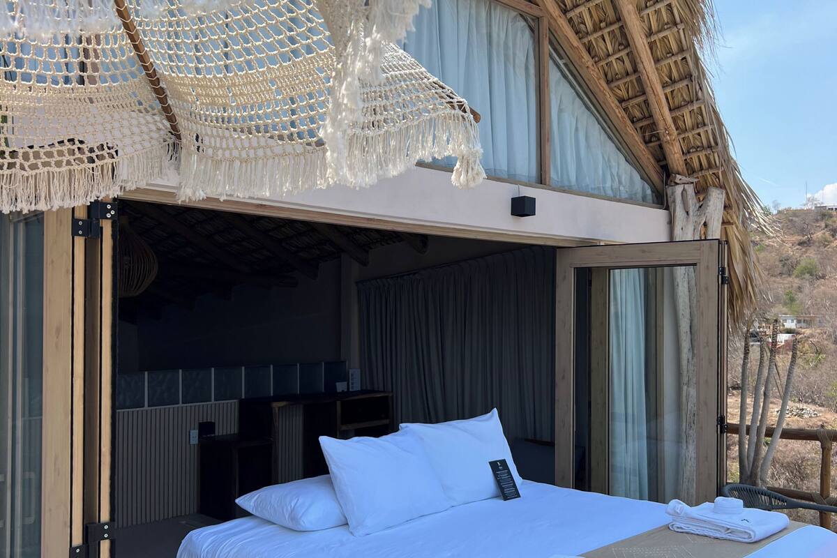 A bed with white linens, a beige throw blanket, and a white towel on a platform bed in a room with a thatched roof and a view of the landscape.