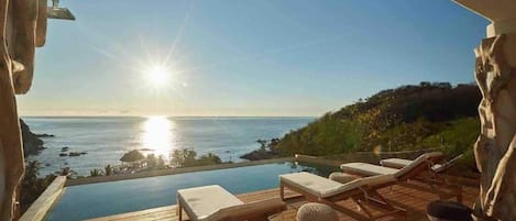 An infinity pool with lounge chairs, a wooden deck, and a view of the ocean at sunset.