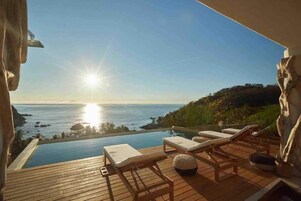 Una piscina infinita con sillones de descanso, una terraza de madera y vistas al océano al atardecer.