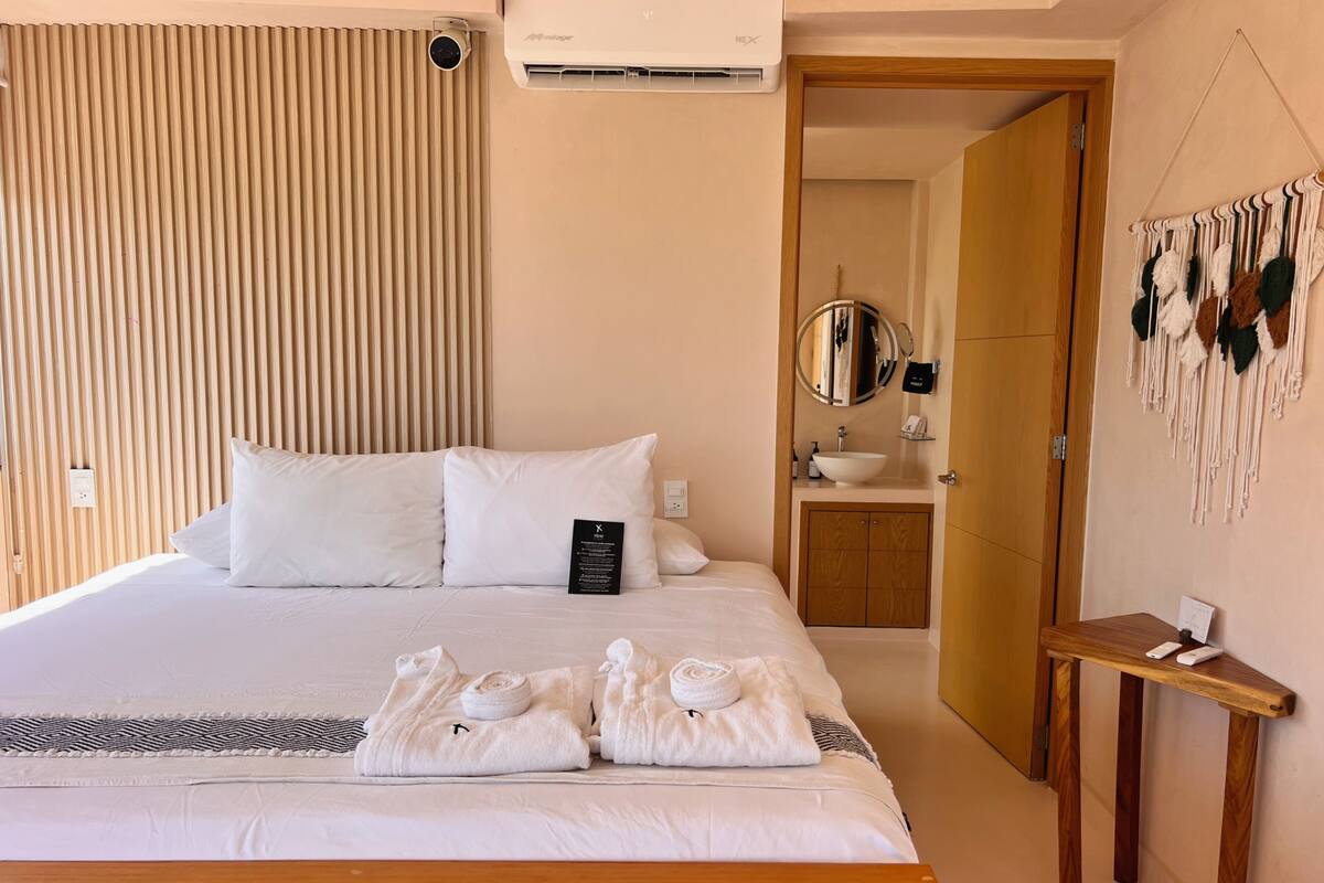 A hotel room with a bed, a wooden bedside table, a mirror, a sink, and a door leading to another room.