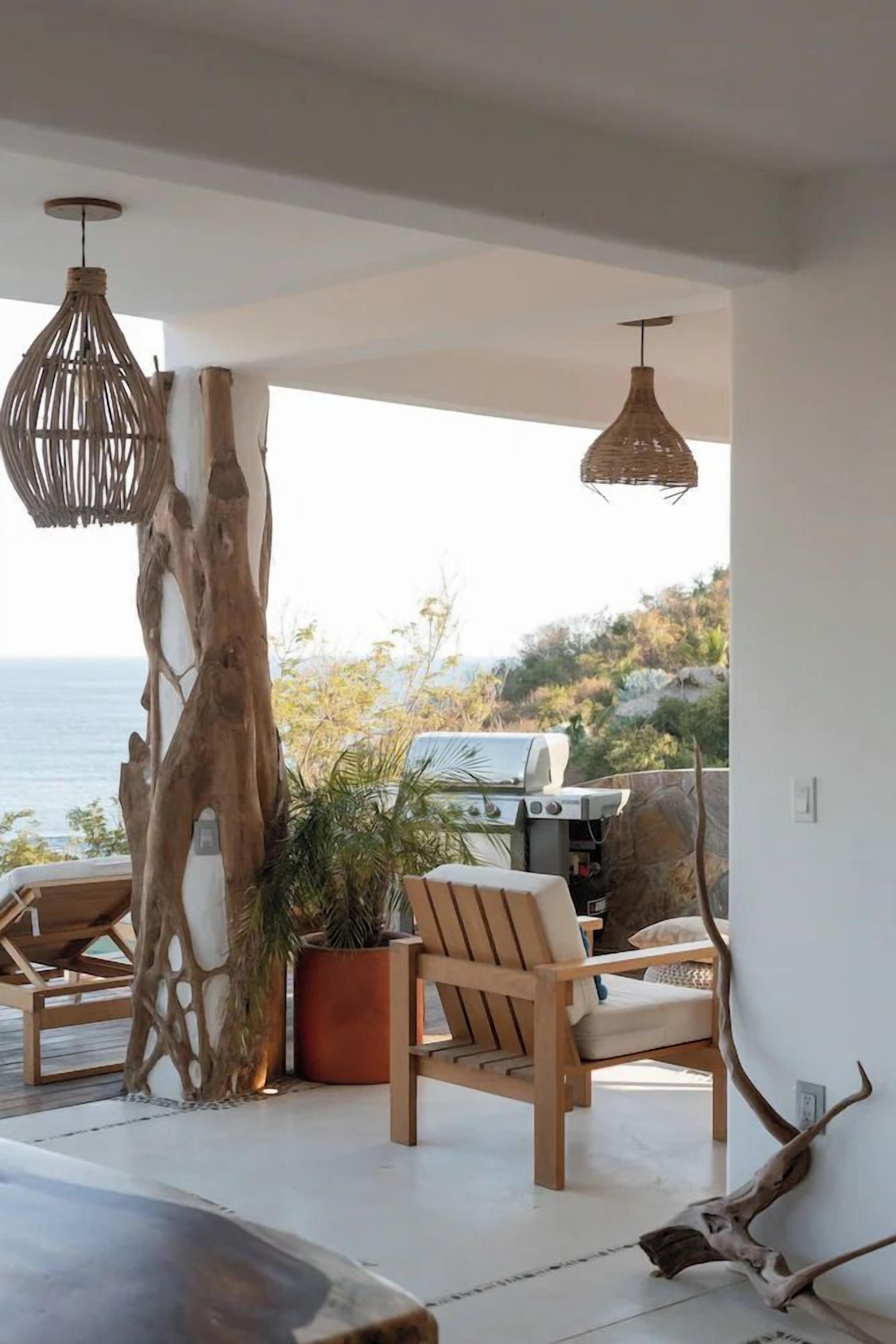 Un balcón con vista al mar, una parrilla, mobiliario de madera y luces colgantes.
