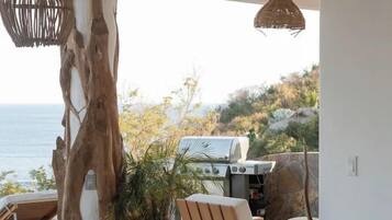 Un balcón con vista al mar, una parrilla, mobiliario de madera y luces colgantes.