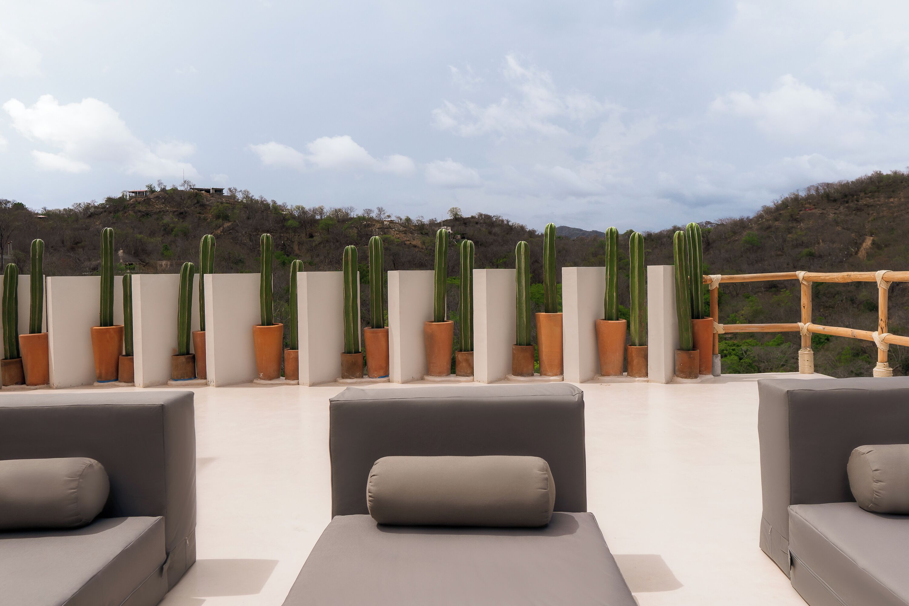 Eine Dachterrasse mit mehreren grauen Sofas und einer Reihe von Kübelkaktussen vor einer Hügelkulisse.