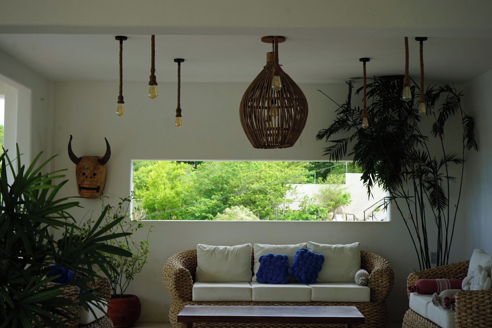 A room with a woven sofa, a wooden mask on the wall, and a window with a view of trees.