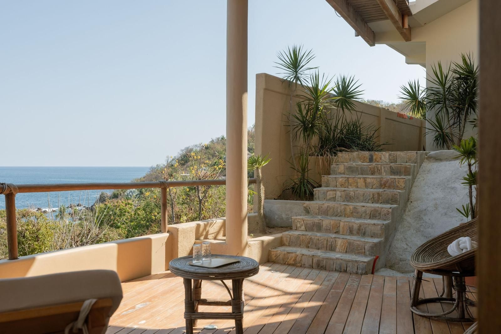 Una terraza de madera con una mesa y sillas, una escalera de piedra y vistas al mar.