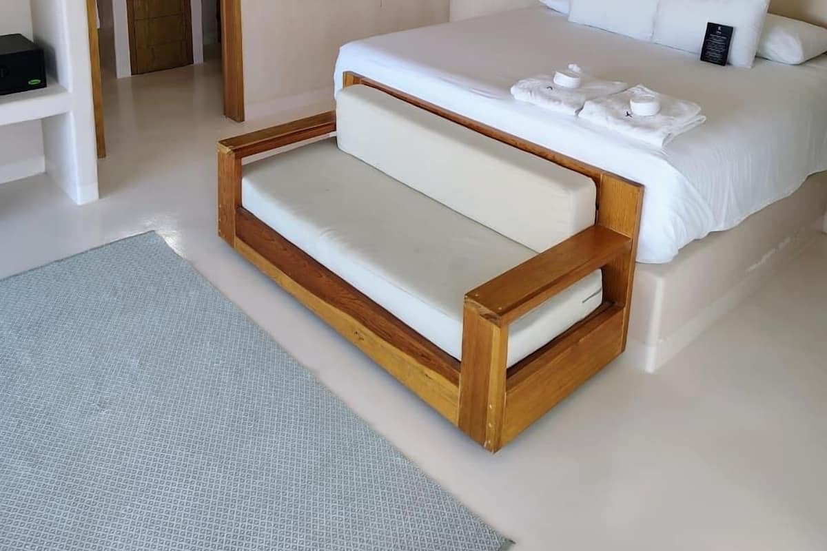 A modern bedroom with a large bed, a wooden bench, a small bathroom, and a ceiling-mounted camera.