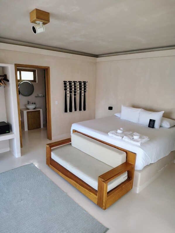 A modern bedroom with a large bed, a wooden bench, a small bathroom, and a ceiling-mounted camera.