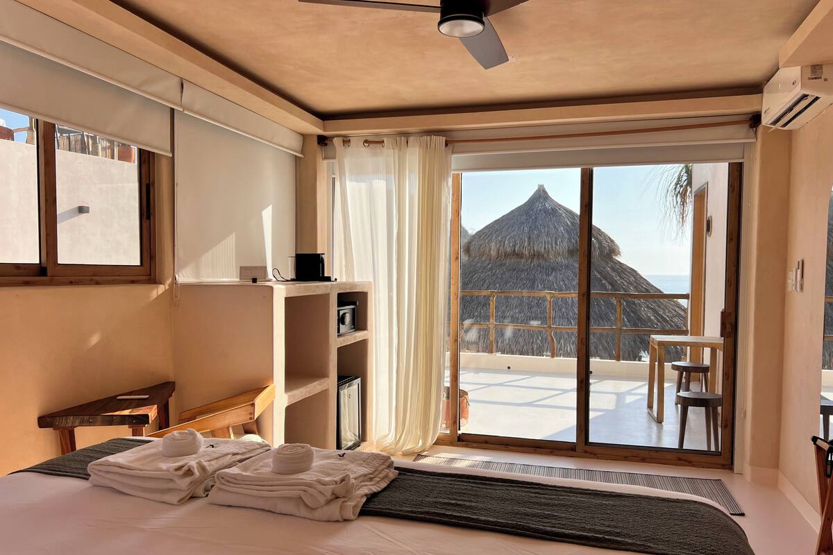 A hotel room with a bed, a desk, a chair, a shelf with a camera, and a view of a thatched-roof structure.