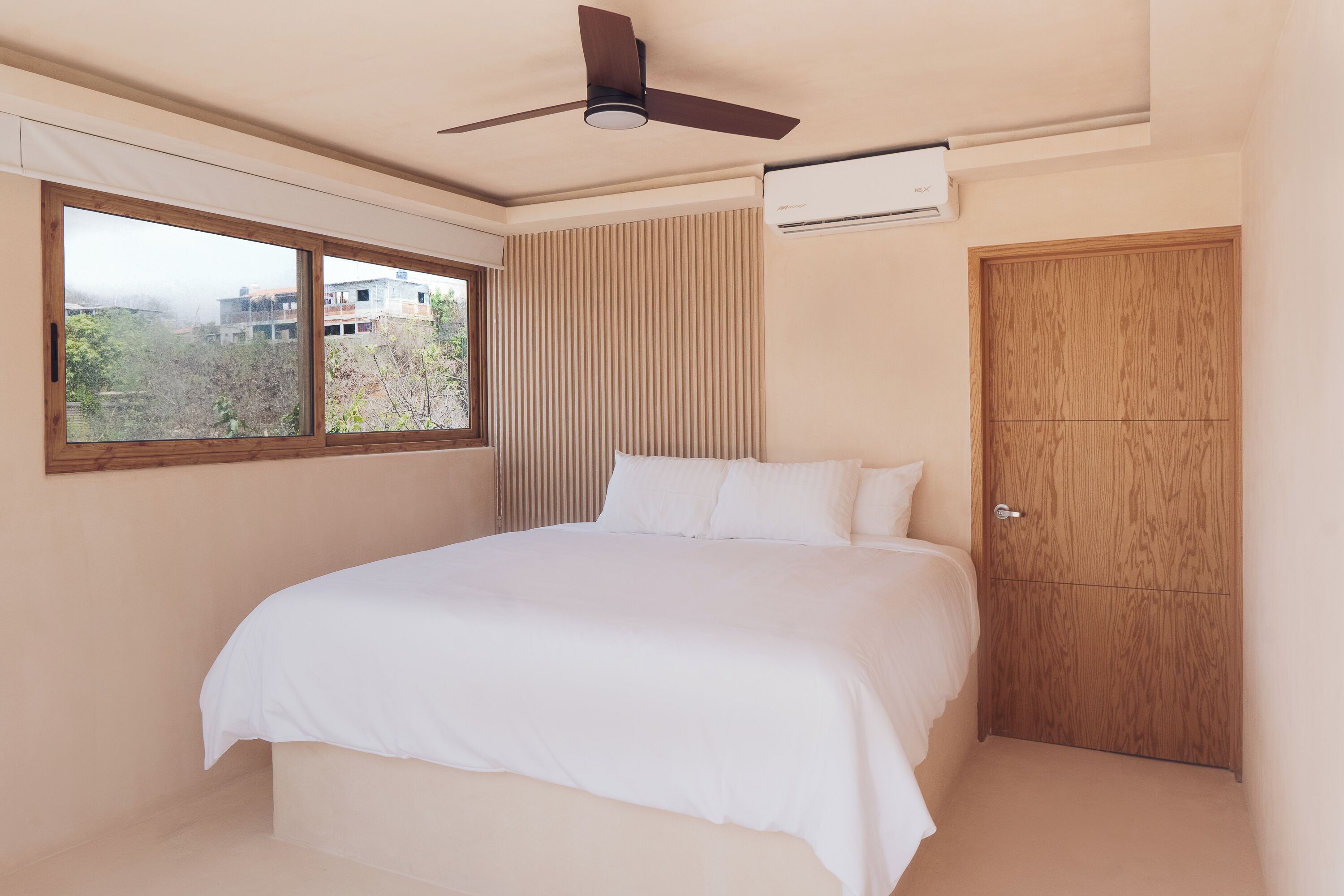 A bedroom with a bed, a ceiling fan, an air conditioning unit, a window, and a wooden door.