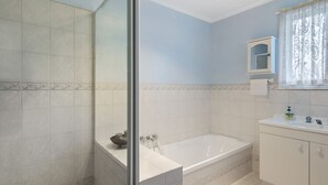Ein Badezimmer mit einer Badewanne, einer Duschkabine aus Glas, einem Waschbecken mit Spiegel und einem Fenster mit floralem Vorhang.