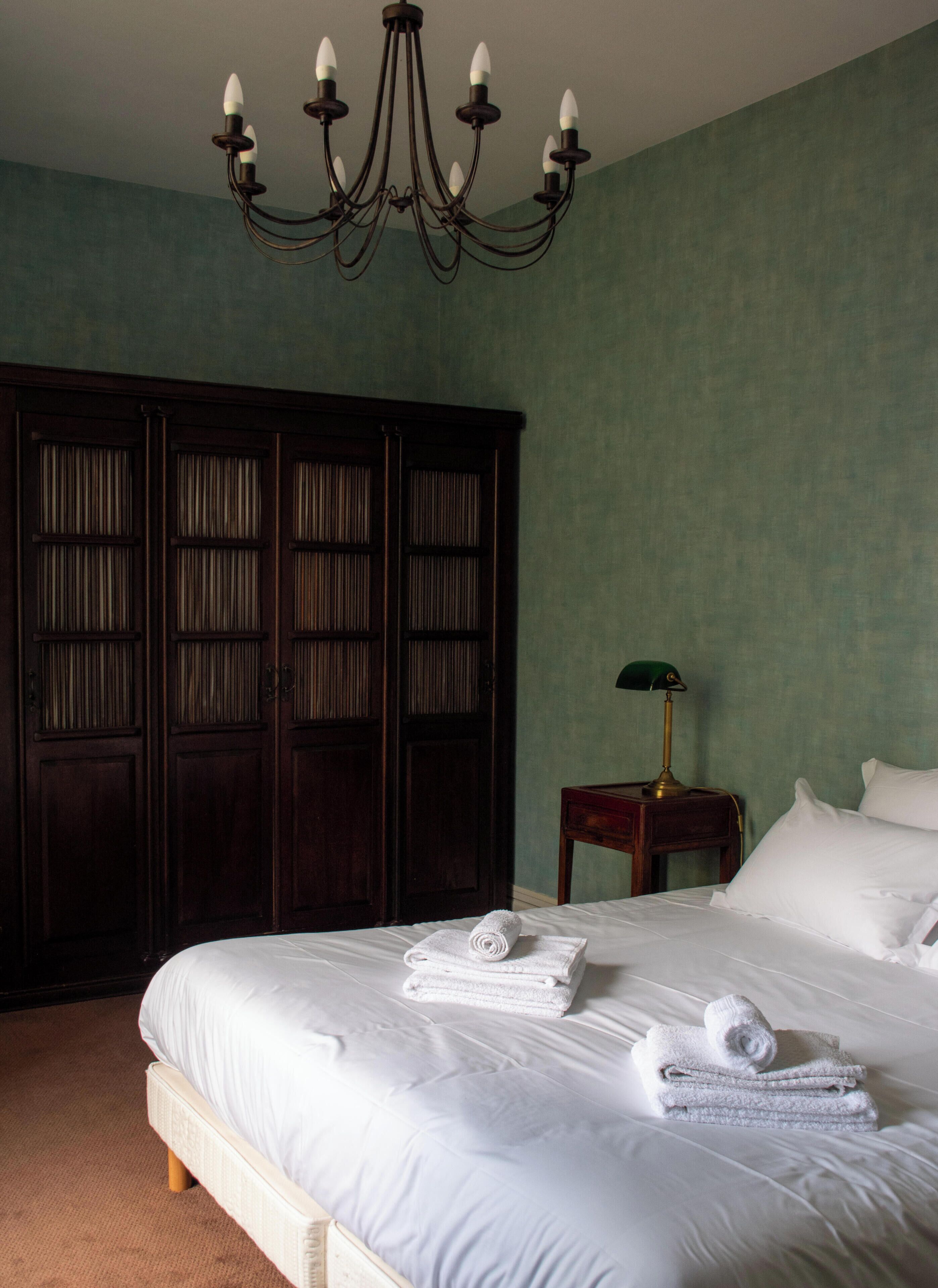 A bedroom with a large bed, a wooden wardrobe, a nightstand with a lamp, and a chandelier.