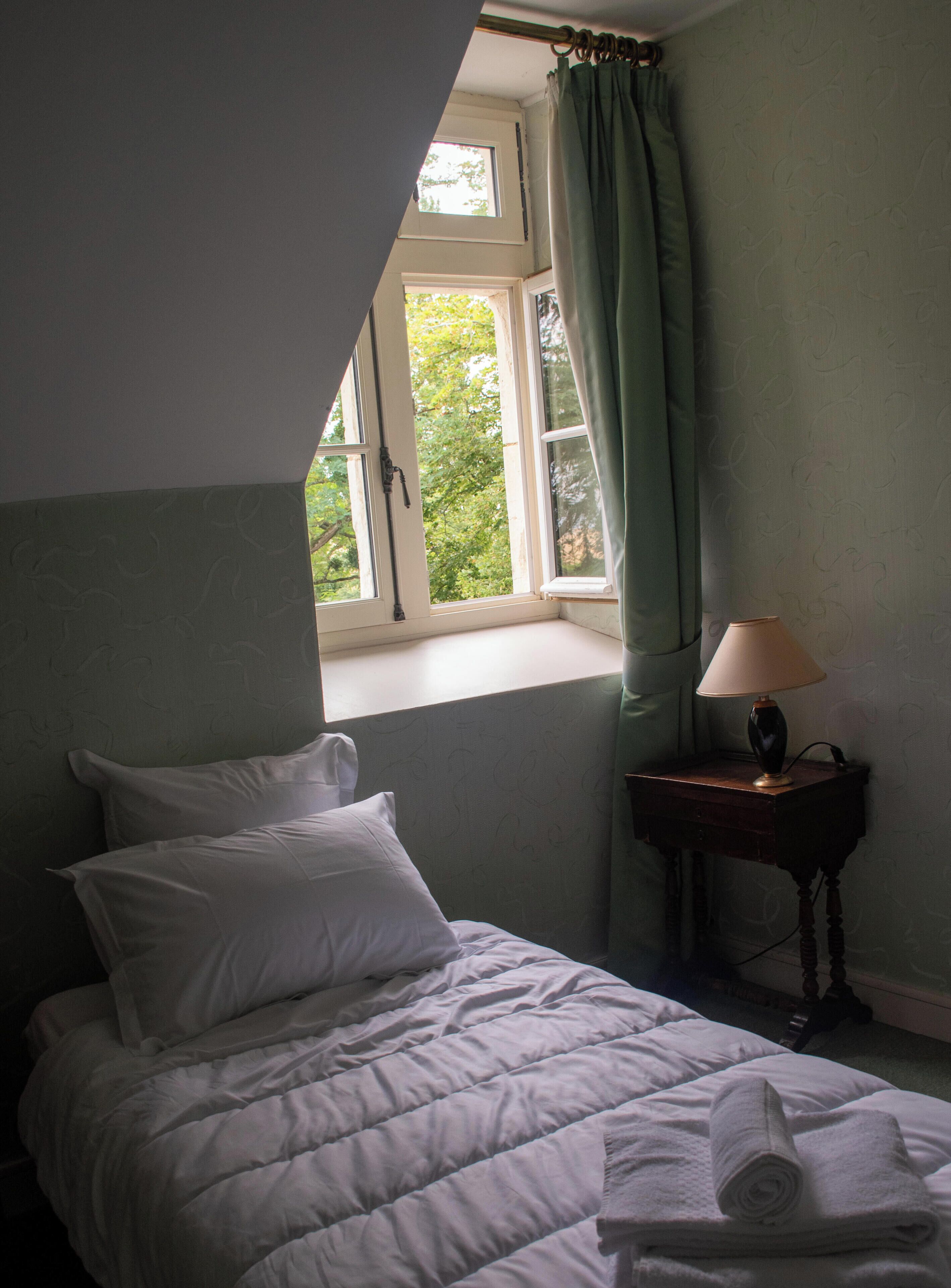 A bedroom with a bed, a window with curtains, a bedside table with a lamp, and a view of greenery outside.