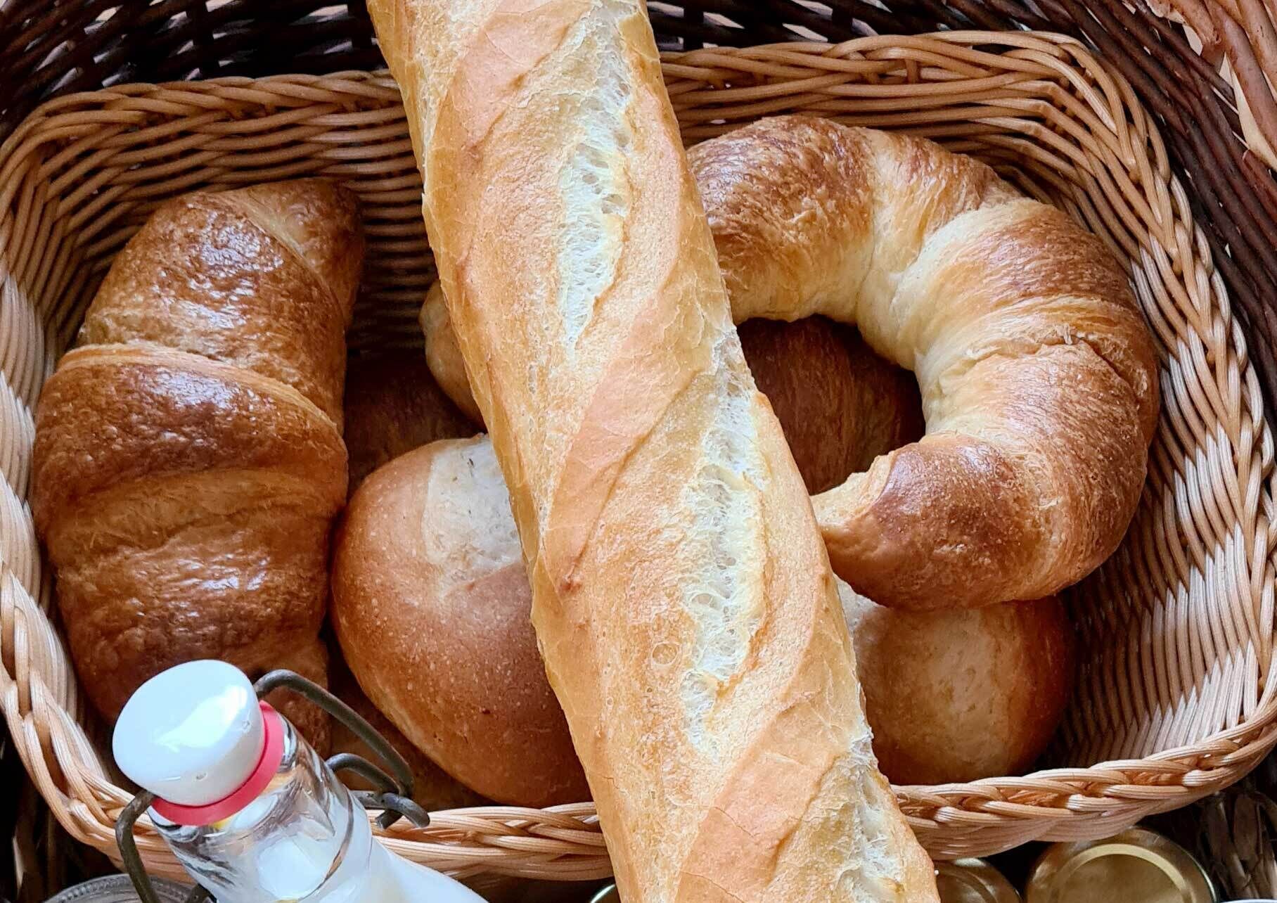 A basket containing various types of bread, including baguettes and rolls, with a bottle of milk and jars of preserves.