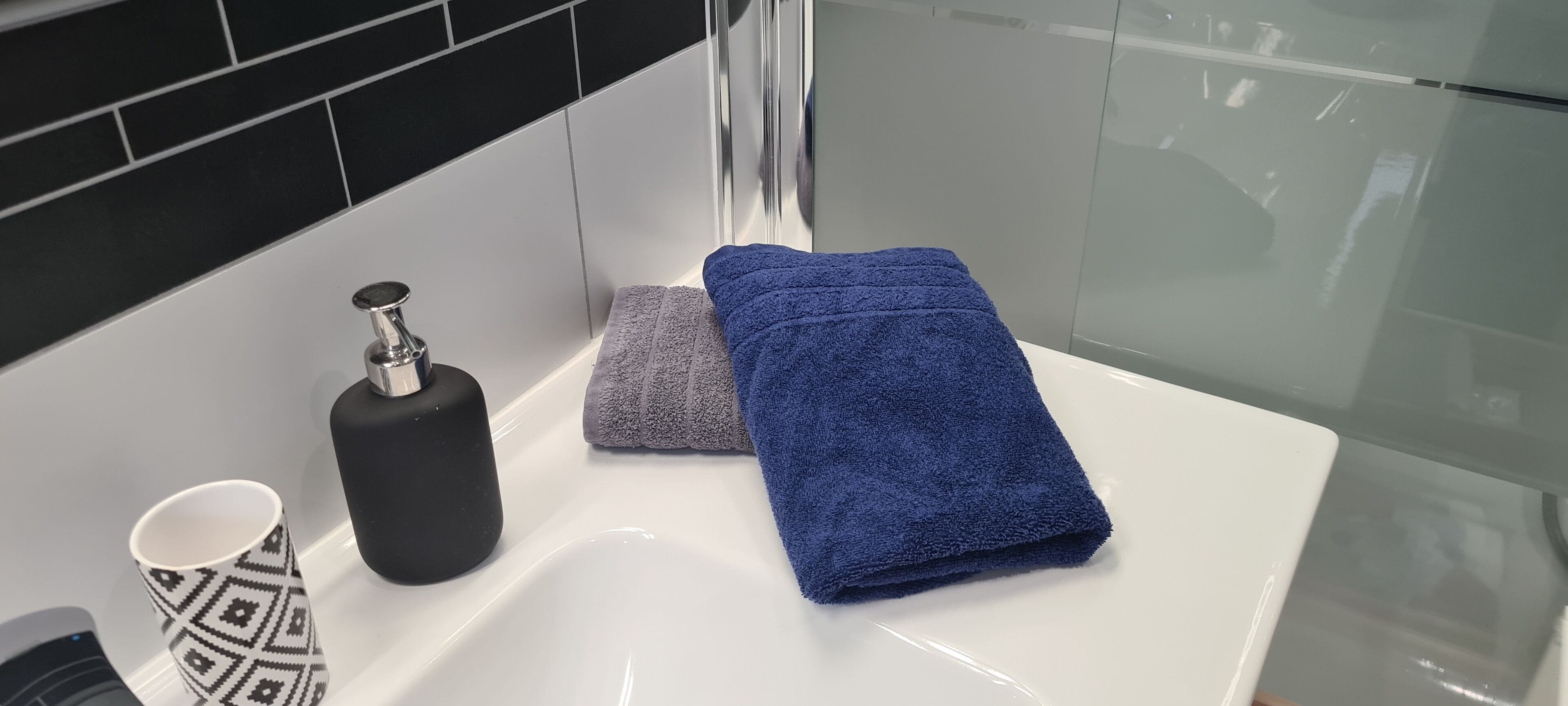 Une salle de bain avec un lavabo blanc, un distributeur de savon noir, une serviette bleue, une serviette grise et une tasse à motifs.