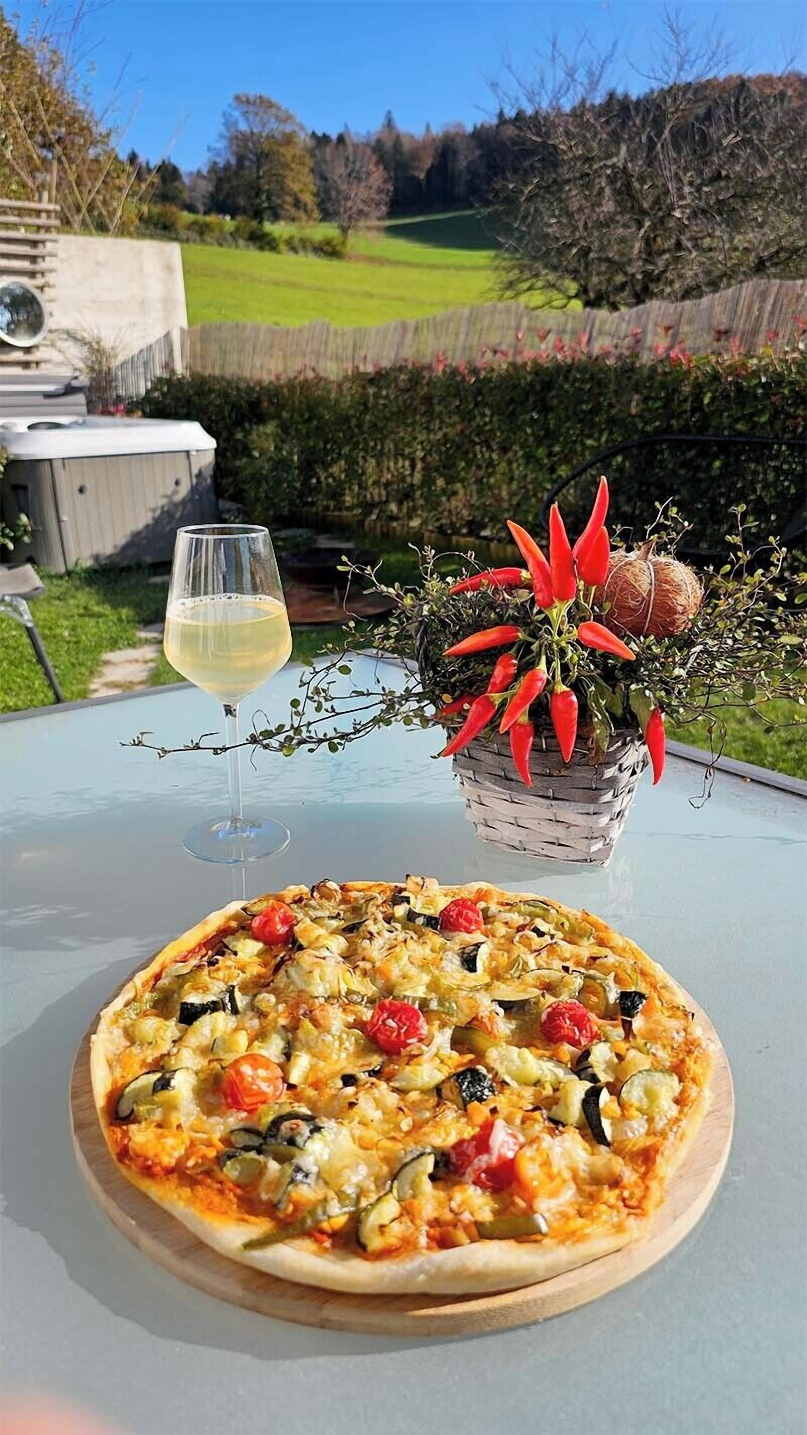 A pizza with various vegetables on a wooden board, a glass of white wine, and a potted plant with red chili peppers on a table outdoors.