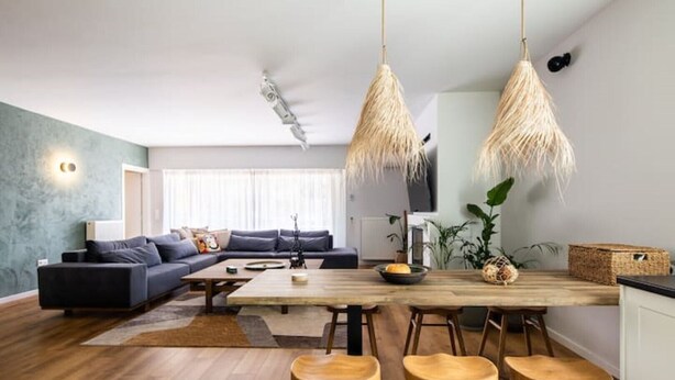 Ein modernes Wohnzimmer mit einem dunkelgrauen Sofa, einem Holzespeisetisch mit Barhocker und einem großen Fenster mit transparenten Vorhängen.