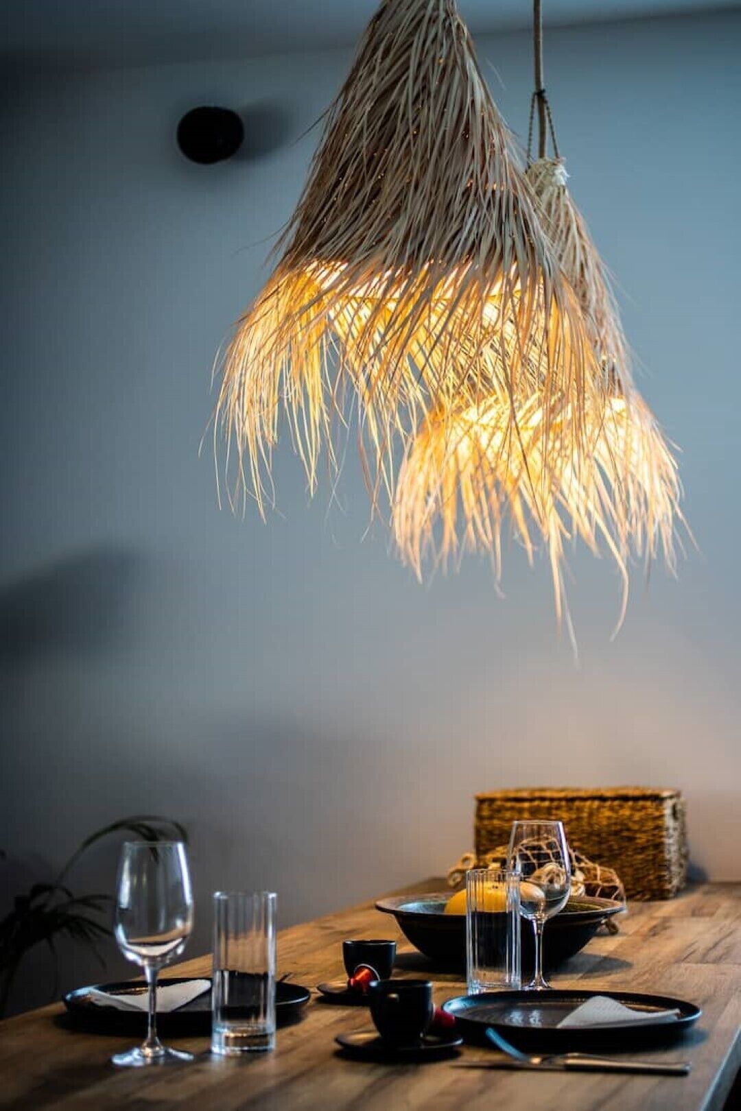 A dining table with a woven pendant light fixture, clear glasses, and a basket of lemons.
