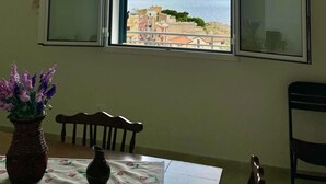 A room with a large window offering a view of the sea and buildings. There is a table with a vase of flowers and a chair. - Stunning apartment only 100 meters from the sea and spectacular gulf view. (Santa Flavia)