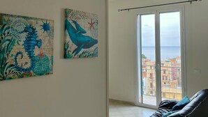 A spacious living room with a black sofa, two wall-mounted artworks depicting sea creatures, and a large window offering a view of the city and sea. - Stunning apartment only 100 meters from the sea and spectacular gulf view. (Santa Flavia)