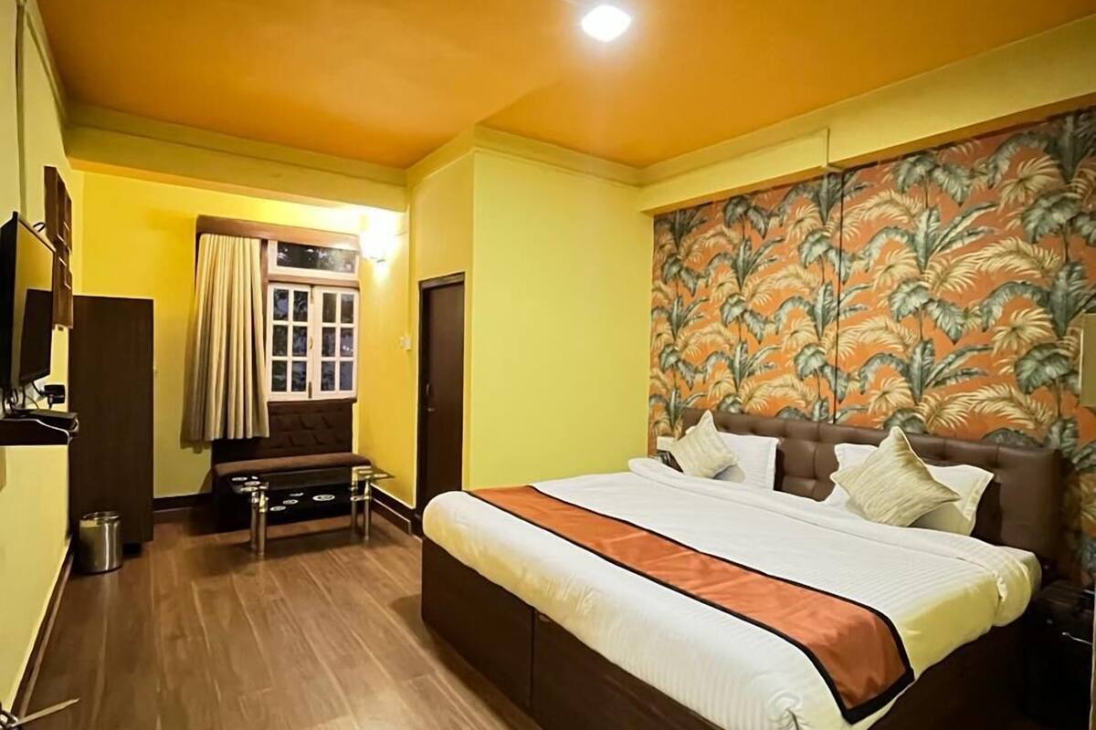 Une chambre avec un grand lit, un plancher en bois, une fenêtre avec des rideaux et un mur orné d’un motif de feuilles tropicales.