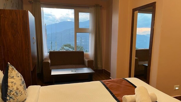 A hotel room with a large bed, a wooden wardrobe, a seating area with a view of mountains, and a mirror reflecting the room. - Little Sikkim Inn (Gangtok)