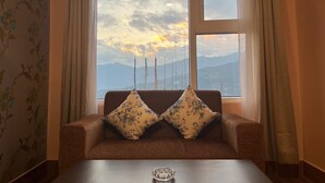 A room with a sofa facing a large window with a view of mountains and a sunset. - Little Sikkim Inn (Gangtok)