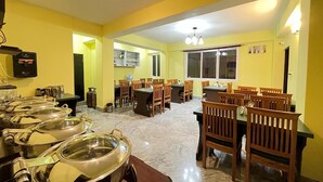 A dining area with multiple tables and chairs, a buffet setup with food containers, and a kitchen in the background. - Little Sikkim Inn (Gangtok)