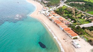 An aerial view of a coastal area with a beach, a row of buildings with red roofs, and a large dark object in the water. - Apartment in Vignola Mare near Sandy Beach (Vignola Mare/Aglientu (SS))
