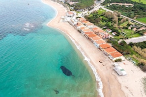 An aerial view of a coastal area with a beach, a row of buildings with red roofs, and a large dark object in the water.