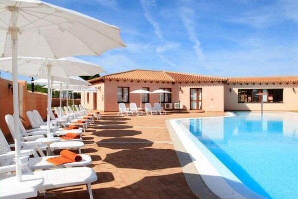 A poolside area with white lounge chairs and umbrellas, a clear blue pool, and a building with a tiled roof.