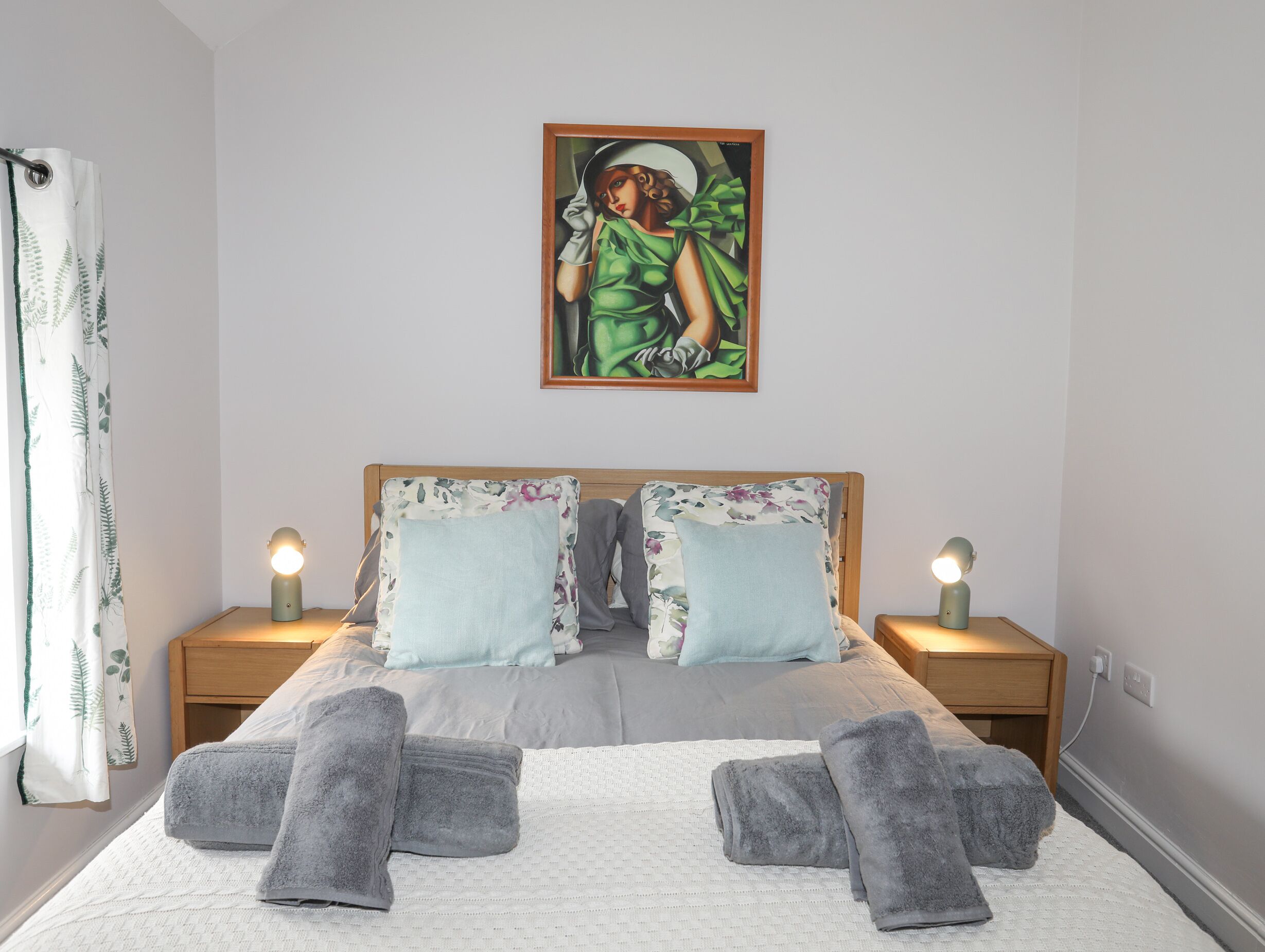 A neatly made bed with two pillows, two bedside lamps, and a painting above it.
