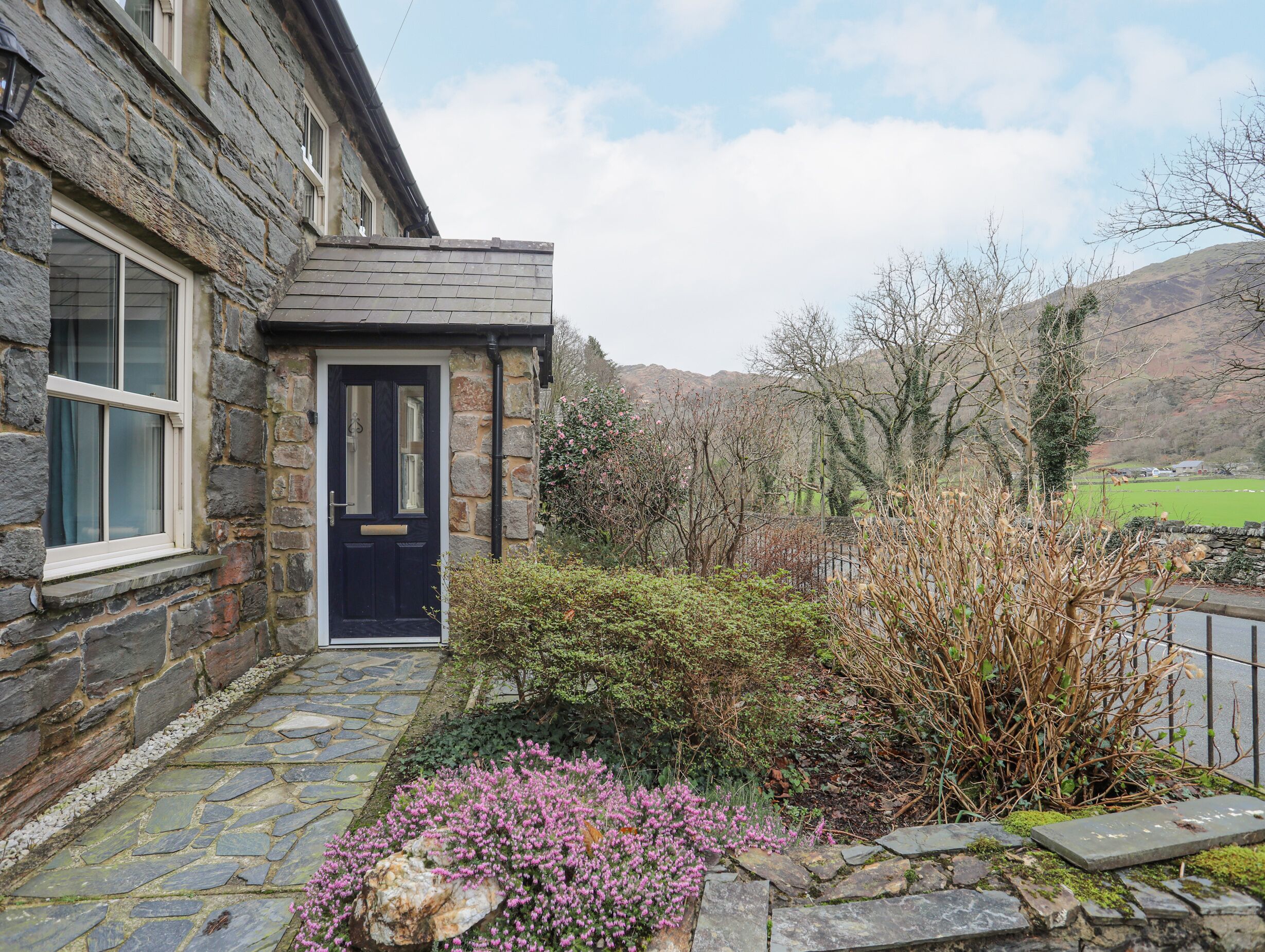 A stone house with a black door, a stone pathway, and a garden with pink flowers.