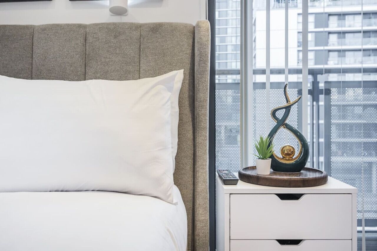 A neatly made bed with white linens, a grey headboard, and a bedside table with a small potted plant and decorative items.