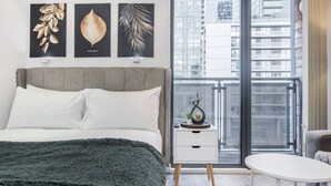 A modern bedroom with a grey upholstered bed, white pillows, and a dark green throw blanket. There are three framed botanical prints on the wall, a small bedside table with a decorative item, and a view of the city through a large window. - The Canary Hub based in Canary Wharf (Greater London)