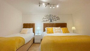 A hotel room with two beds, each with a wooden headboard and yellow bedding. There is a nightstand with a lamp between the beds, and a wall-mounted light fixture above. - Kings Cross St Pancras Oasis (Greater London)