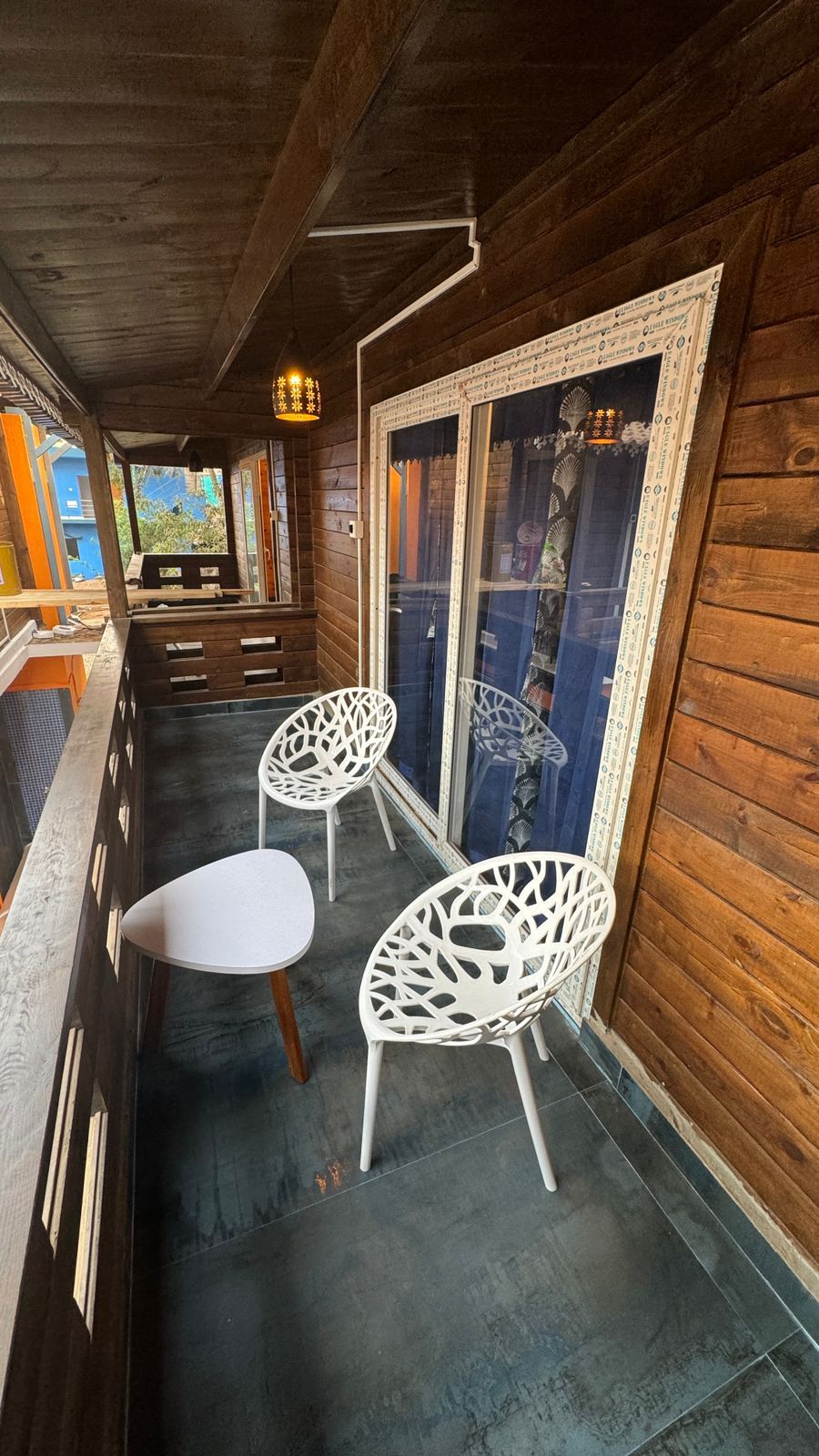 A wooden balcony with two white chairs and a table, a patterned window, and a view of the outside.