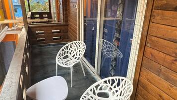 A wooden balcony with two white chairs and a table, a patterned window, and a view of the outside.