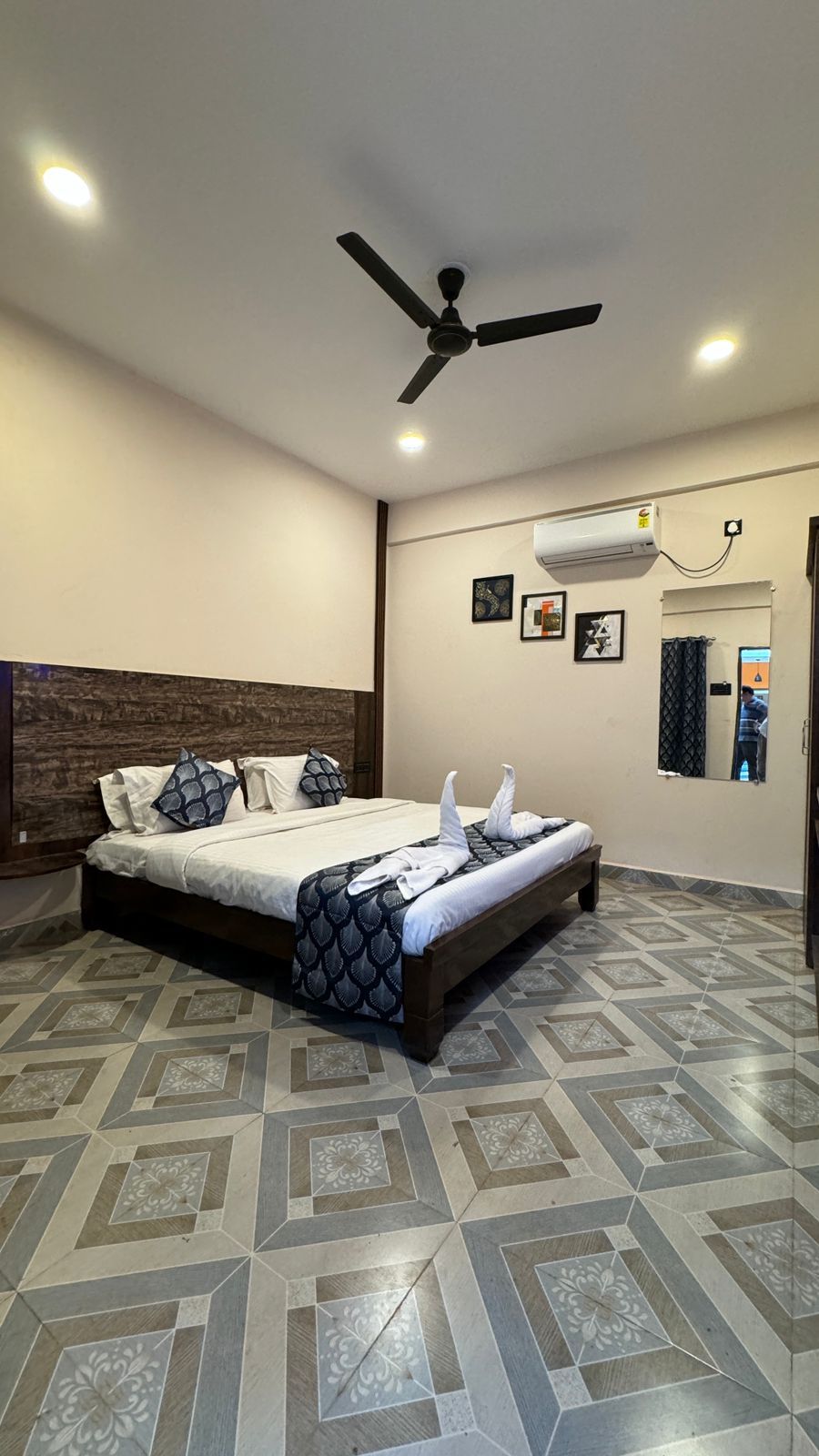 A hotel room with a bed, ceiling fan, air conditioning unit, and patterned floor tiles.