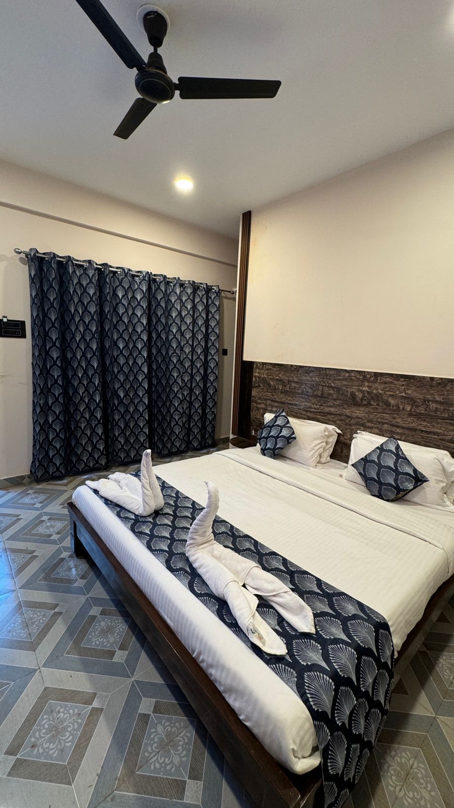 A hotel room with a bed, curtains, a ceiling fan, and a patterned floor.