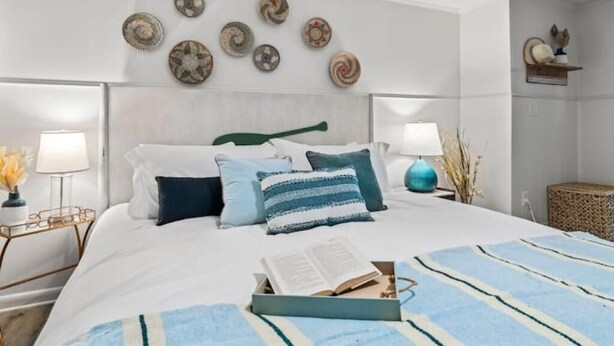 A neatly arranged bedroom with a bed, bedside lamps, decorative plates on the wall, and a small shelf with a statue. - Jax Bch Home 3/2 w/Pets (Jacksonville Beach)