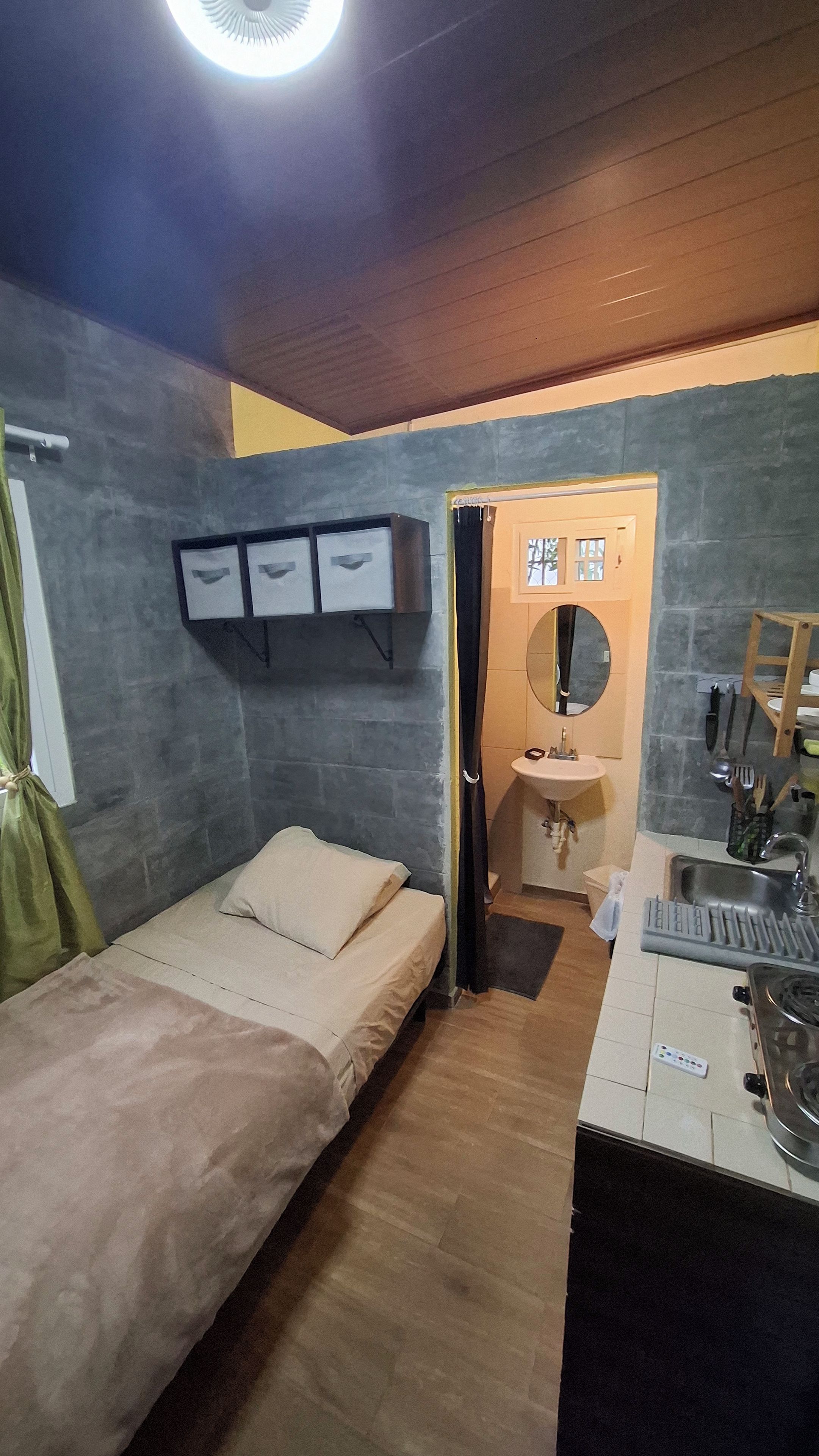 A compact bedroom with a bed, a bedside table, a small bathroom with a sink, and a mirror.