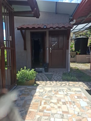 Une maison avec un hall d’entrée carrelé, une porte en bois et un petit jardin agrémenté de plantes.
